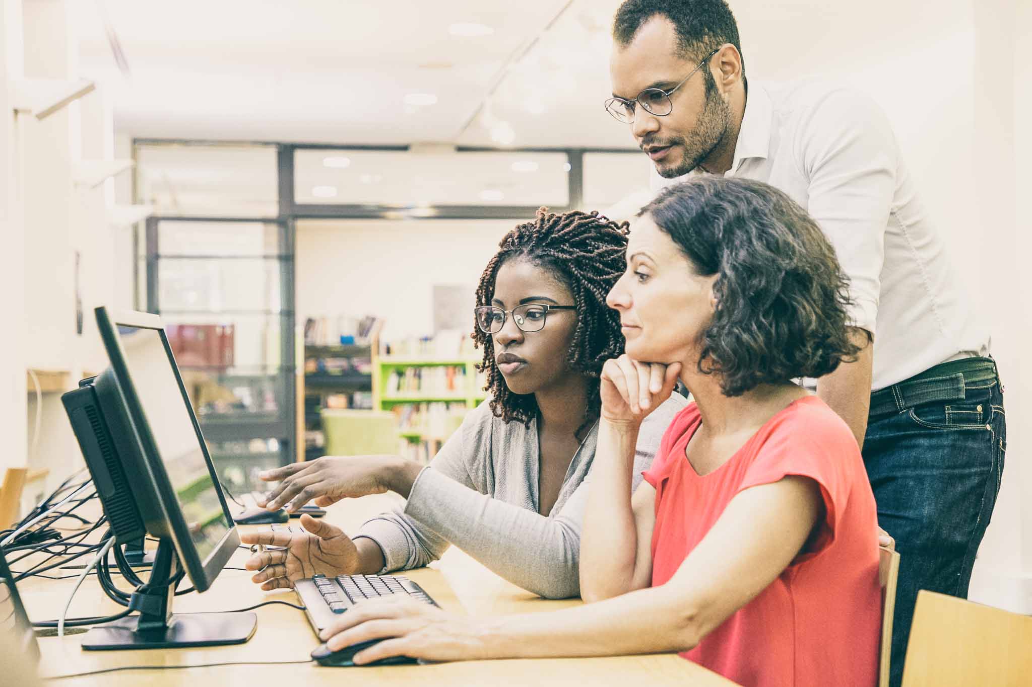 Colleagues huddle over a library computer to share ideas, whispering so they don't disturb others.