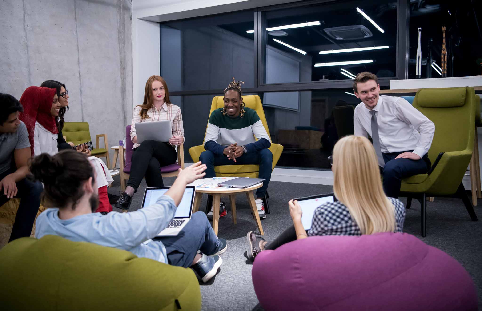 A jovial and enthusiastic team sit around a coffee table, laptops open, and discuss strategy. 