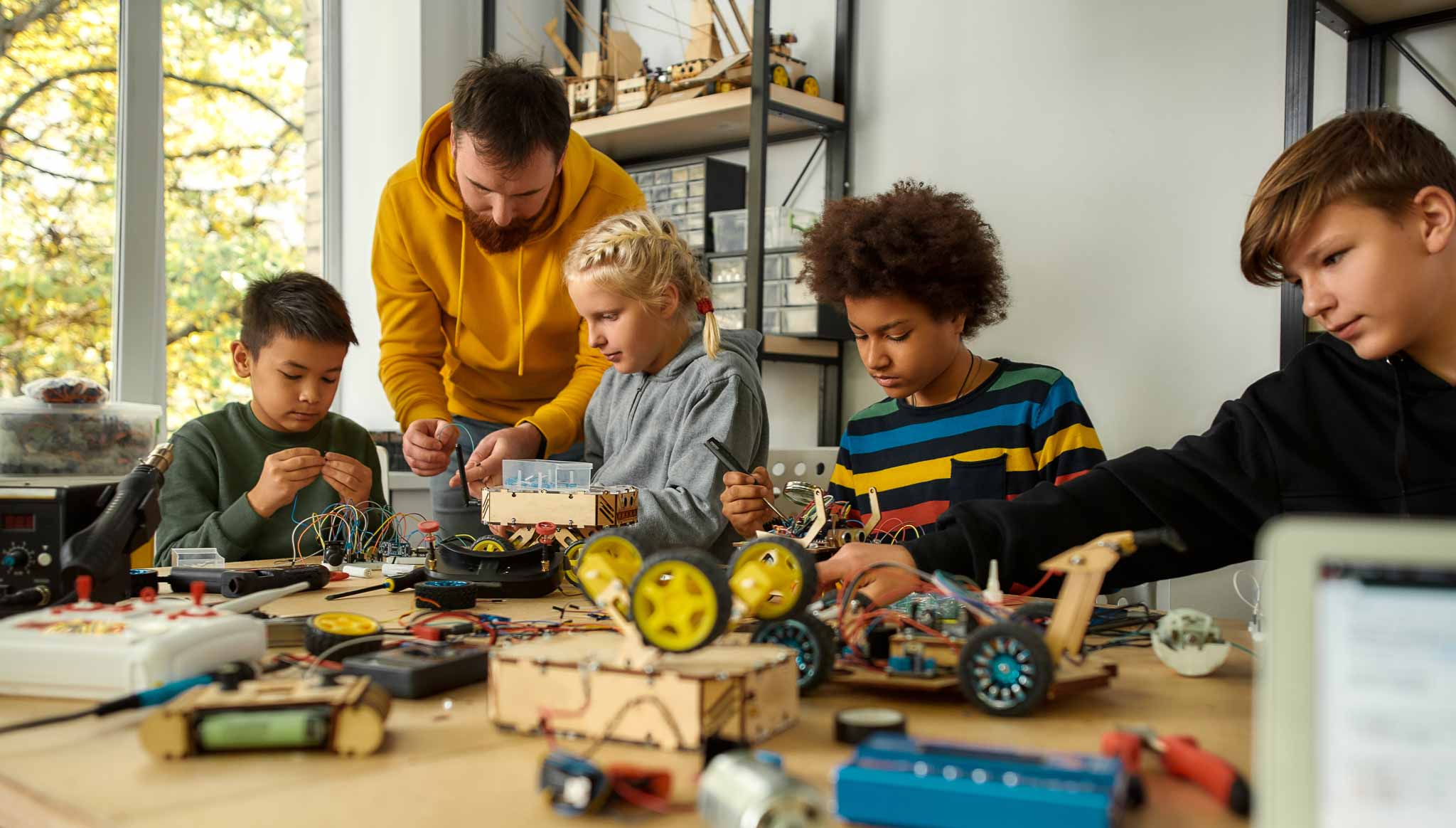 Four children and an adult sit around a colorful table table full of small vehicle components. Some use tools, other are working with adhesives to make small robots.