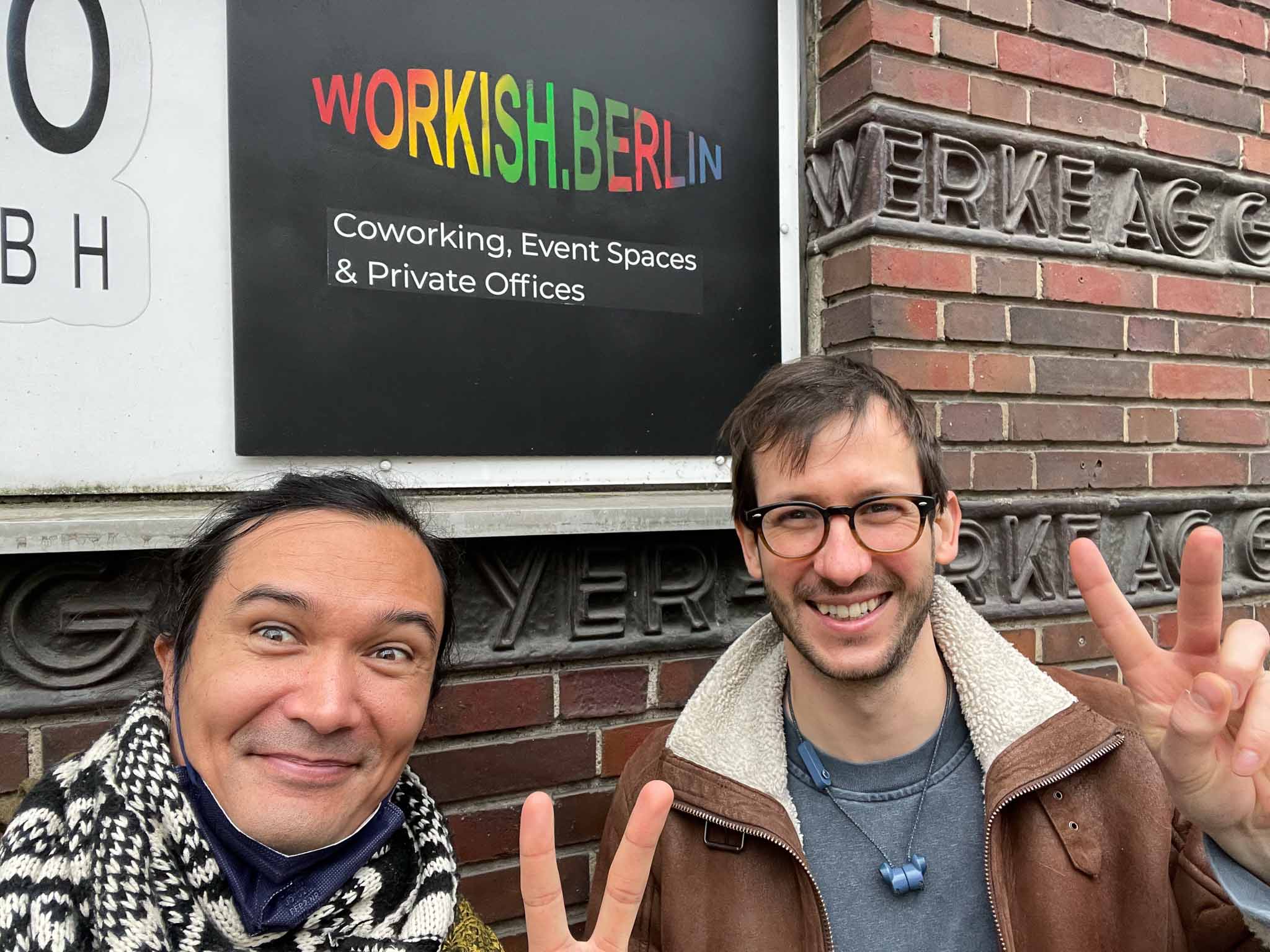 Cobot’s Morgan Sully and Lenny Leiter stand at the entrance to workish.berlin, smiling at the camera and holding up peace signs in front of the colorful workish logo.