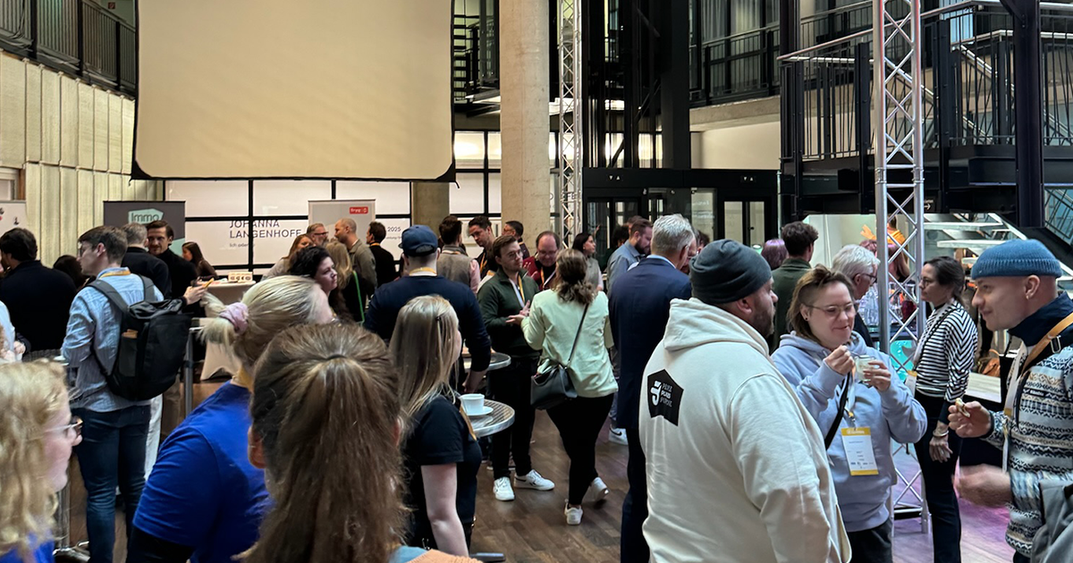 A lively networking session at ZukunftCoworking 2024, with attendees gathered in the main hall, conversing and engaging. People stand in small groups, fostering connections, while the event backdrop and promotional stands are visible in the background.