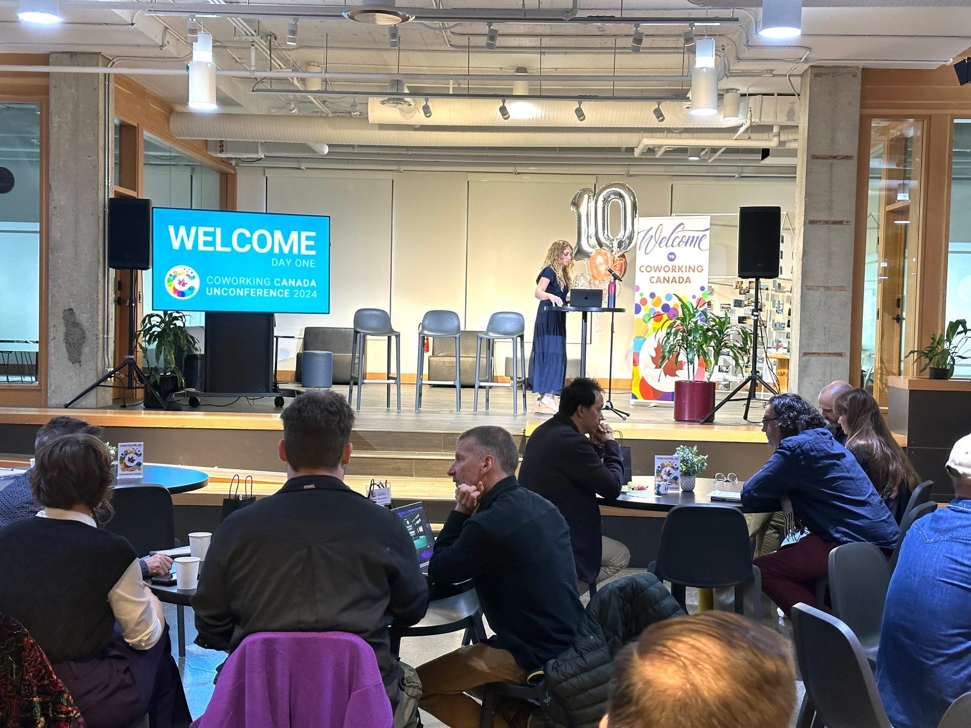 A welcoming scene at Coworking Canada Unconference 2024, Day One. Attendees are seated, engaging in conversations, while the stage is set with a welcome sign, balloons celebrating 10 years of Coworking Canada, and a presenter preparing for the session.