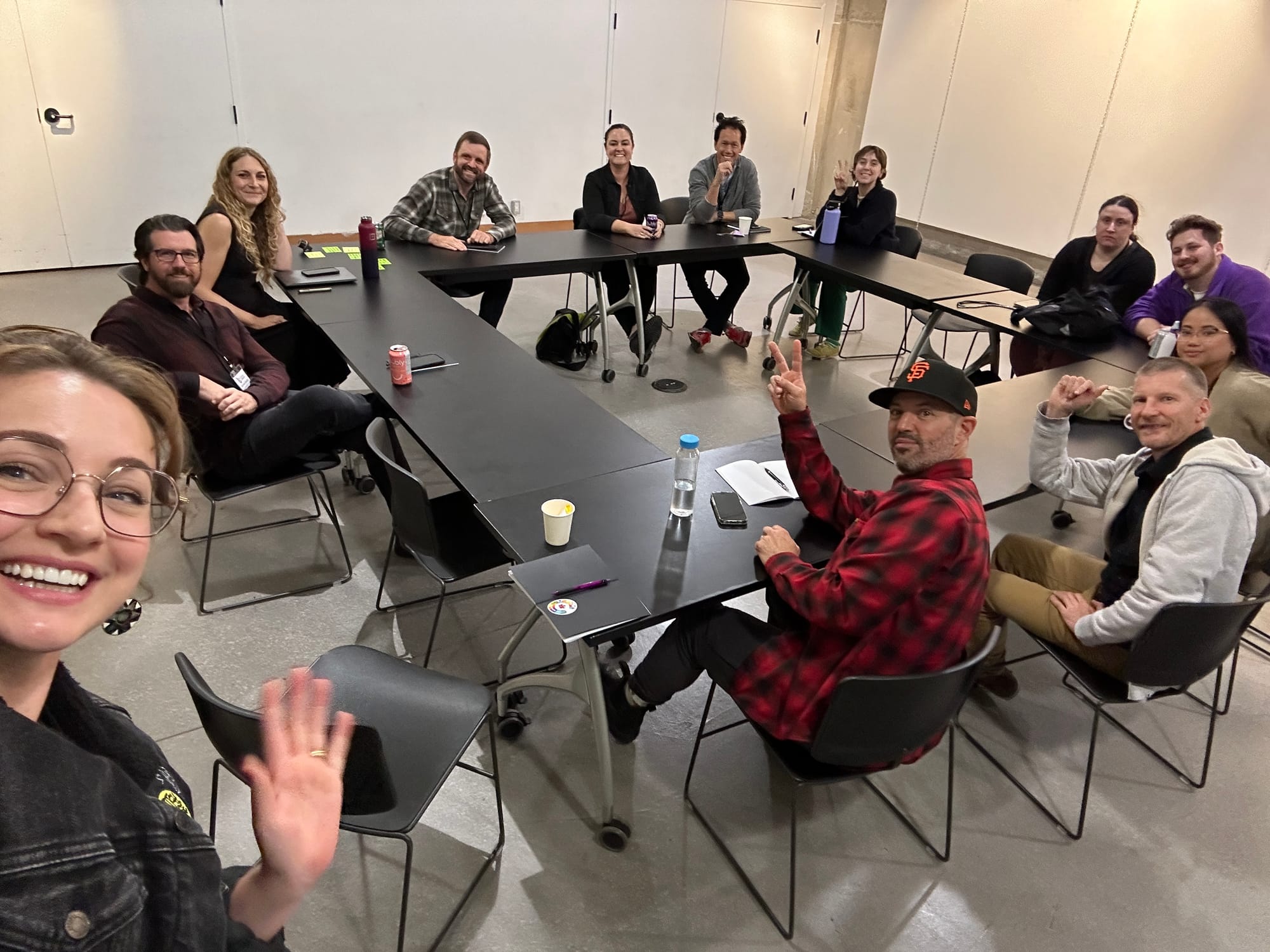 A group of attendees and organizers from Coworking Canada Unconference 2024 gathered around a meeting table, smiling and engaging in discussions. One person in the foreground takes a selfie to capture the moment.